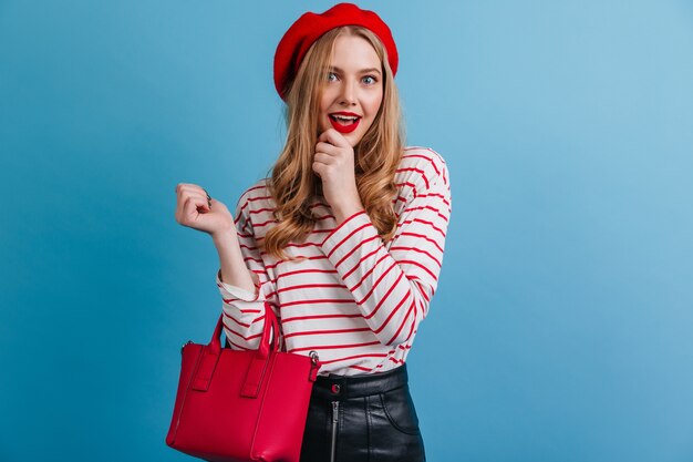 Enchanting girl in red beret. Front view of french blonde lady isolated on blue wall.