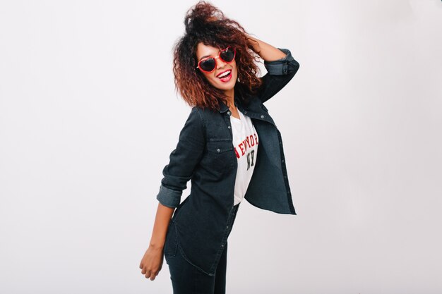 Enchanting girl in red sunglasses posing with hand up. Laughing fashionable female model with african hairstyle enjoying.