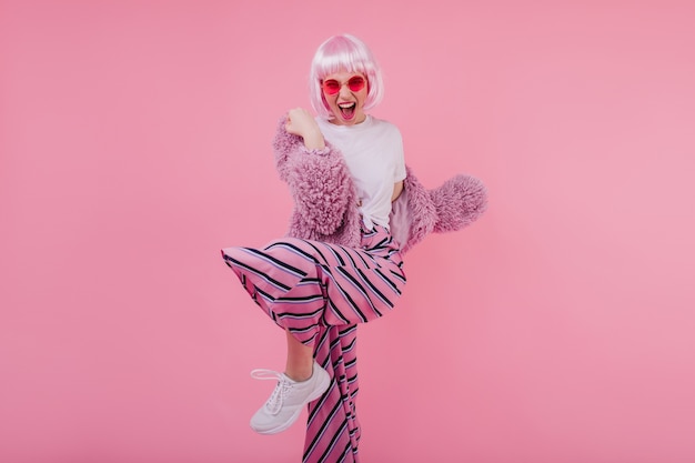 춤을 추는 동안 긍정적 인 감정을 표현하는 세련된 흰색 운동화에 매혹적인 소녀. 모피 재킷에 포즈를 취하는 분홍색 머리를 가진 웃는 아가씨