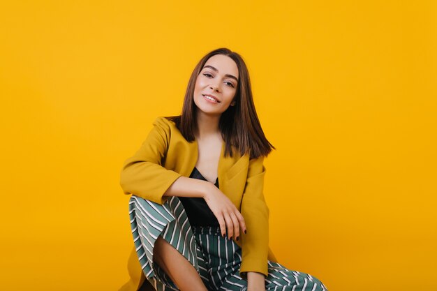 enchanting girl in trendy jacket sitting on the floor. romantic young lady with brown hair posing with interedted smile.