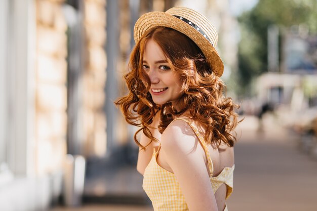 Enchanting girl with dark curly hair fooling around outdoor in warm summer day. Amazing ginger female model in hat and yellow dress laughing on urban street.