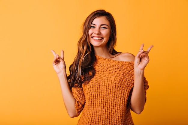 Enchanting girl with long hairstyle posing with peace sign on orange. Glad female model in trendy knitted sweater laughing in studio.