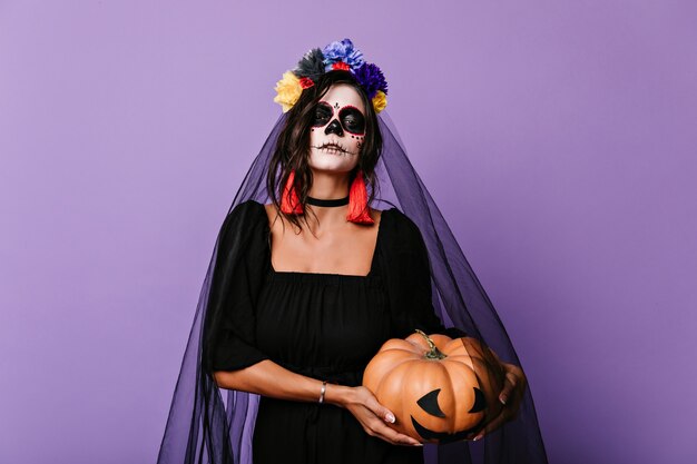 Enchanting girl with scary mexican makeup preparing for halloween . Indoor shot of romantic dead bride in black veil holding pumpkin.