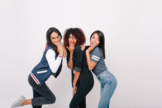 Enchanting international female friends in sport attires posing together. curly mulatto girl in black outfit joking with university mates.