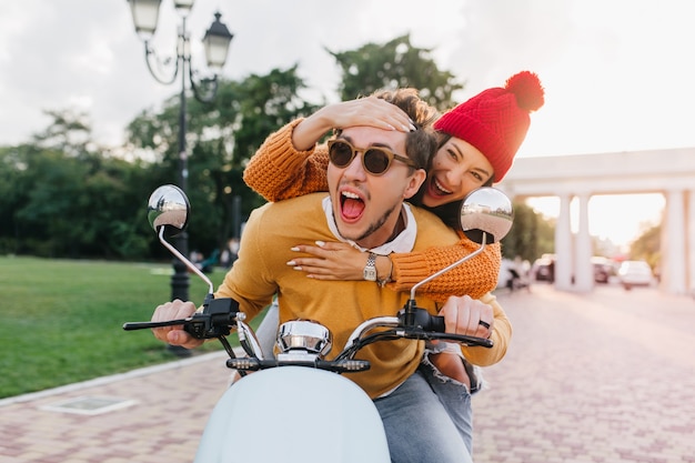 Foto gratuita incantevole signora in cappello rosso che tocca la fronte del ragazzo con un sorriso mentre guida lo scooter