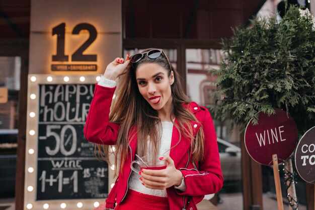 Enchanting lady in red jacket posing with tongue out on the street