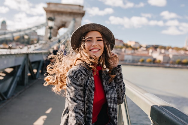 Enchanting lady with gorgeous curly hair posing with pleasure on bridge in windy day