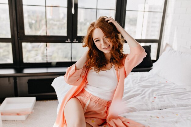 彼女の巻き毛で遊んでいる魅惑的な笑顔の女の子。ベッドに座ってパジャマで熱狂的な女性モデルを笑う。