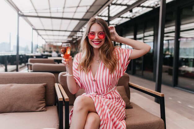 Enchanting white girl in pink sunglasses posing with wineglass in hand. Laughing charming woman in striped dress relaxing in cafe.