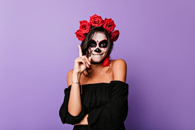Enchanting woman with roses in black hair waiting for halloween party. Happy latin female model with vampire face painting smiling