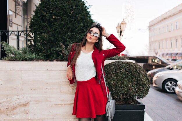 Enchanting young woman in red skirt standing on the street with gray bag