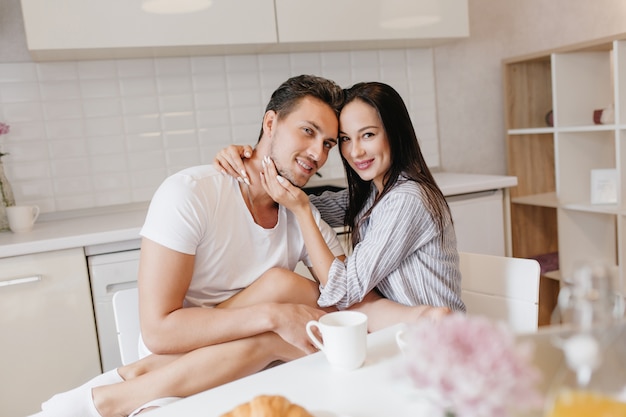 Enchanting young woman sitting on boyfriend's knees while he drinks coffee