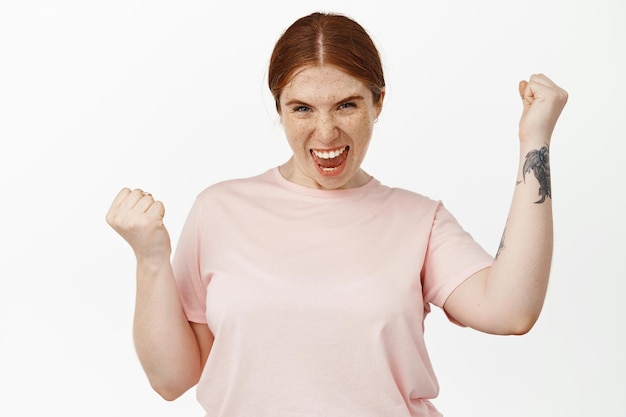 Encouraged and determined redhead girl shouting, yelling with confidence and fist pump, rejoicing over success, achieve goal, celebrating victory, standing against white background