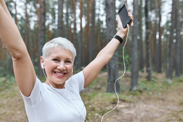 Energetic cheerful retired female with fit slim body posing outdoors in earphones, raising hands, holding cell phone
