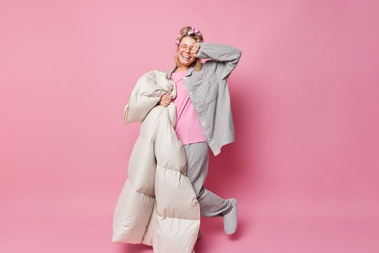 Free Photo energetic cheerful woman wears slumber suit and slippers makes hair curly applies rollers holds soft blanket isolated over pink background going to have healthy sleep. people and rest concept