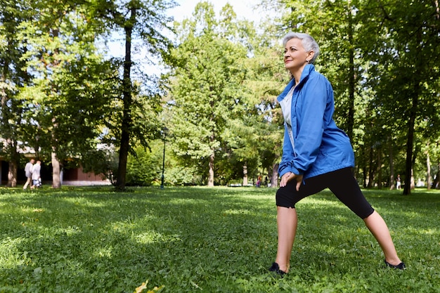 Energetic female pensioner in stylish sportswear choosing healthy active lifestyle training on green grass in forest or park, doing lunges, having happy joyful look. Aged people, fitness and summer