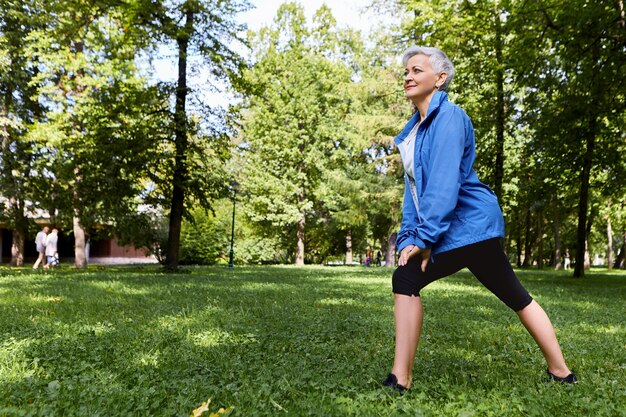 Энергичная пенсионерка в стильной спортивной одежде выбирает тренировки здорового активного образа жизни на зеленой траве в лесу или парке, делает выпады, имея счастливый радостный вид. Пожилые люди, фитнес и лето