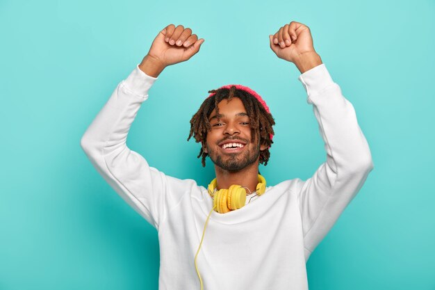 Energetic guy has dread hair, lifts hands up, dances lively and smiles broadly, wears white jumper, headphones on neck, being entertained at party
