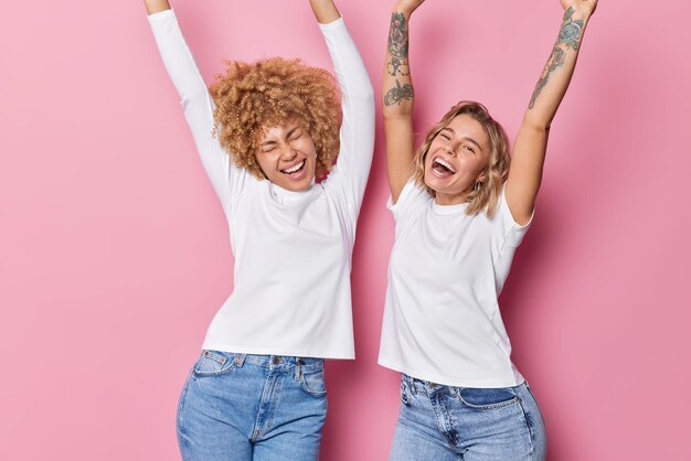 Energetic happy women dance happily keep arms raised up celebrate something enjoy partying express positive emotions move against pink background have fun Cheerful friends have upbeat mood