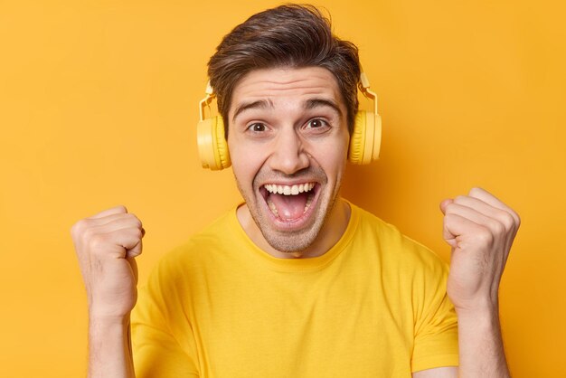 활력이 넘치는 긍정적인 성인 남성이 주먹을 꼭 쥔 채 승자나 챔피언이 귀에 스테레오 헤드폰을 착용하고 행복으로 가득 차서 노란색 배경에서 격리된 좋아하는 음악을 듣는 것을 즐깁니다.