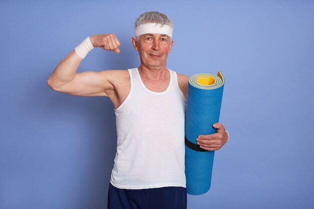 エネルギッシュな年配の男性は、体育をし、ヨガマットを持って、上腕二頭筋と彼の力を示しています