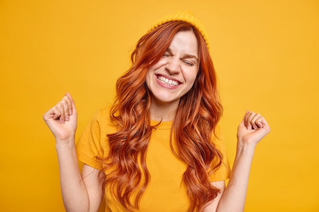 Energetic upbeat millennial girl smiles happily clenches fists from joy celebrates successful day has natural red wavy hair wears hat casual t shirt 