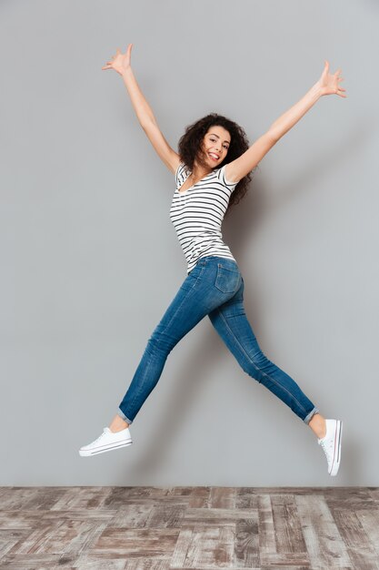 Energetic woman 20s in striped t-shirt and jeans jumping with hands throwing up in air over grey