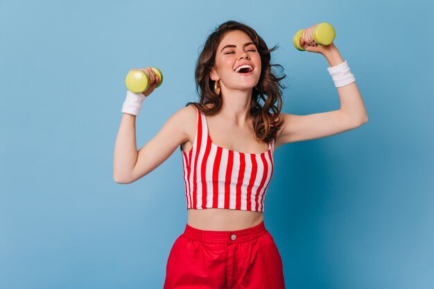 Energetic woman in clothes in style of 80s laughs and does exercises for hands with dumbbells on blue wall