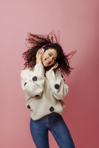 Energetic young caucasian brunette girl listens to music in headphones and dances on pink background Fluffy curlyhaired lady in sweater and jeans spends time alone Good mood concept