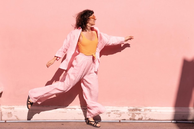 Бесплатное фото Энергичная молодая кавказская женщина в свободной одежде движется летящей походкой вдоль розовой стены в солнечный день