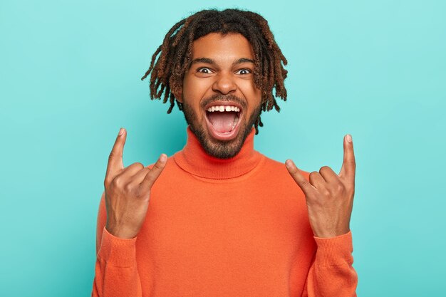 Energized dark skinned young man rocks on party, brings positive vibes, shows rock n roll gesture, keeps mouth opened, has dreadlocks, wears orange turtleneck, isolated over blue background.