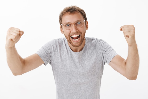 Energized excited and enthusiastic team leader in glasses and grey t-shirt raising fists in cheer yelling from supporting and encouraging workers standing amused over white wall