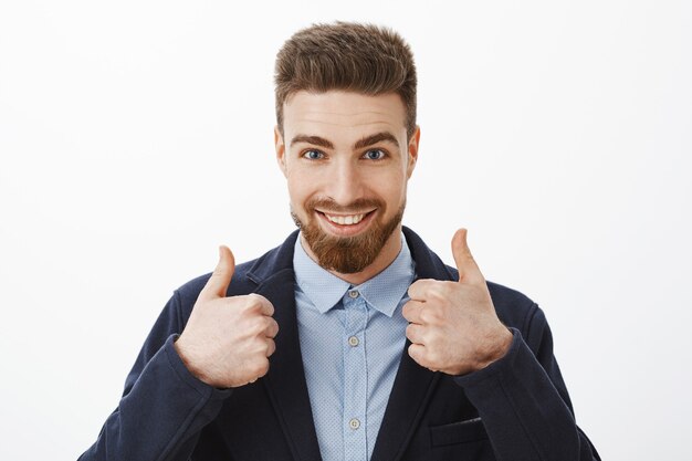 Energized good-looking and assured successful man with beard and blue eyes in stylish formal suit showing thumbs up being assured plan work great standing self-assured against gray wall