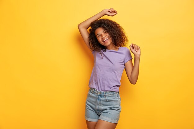 Energized happy Afro American girl raises hands joyfully, being in high spirit, dances to favourite music, has slim figure, dressed in casual clothes