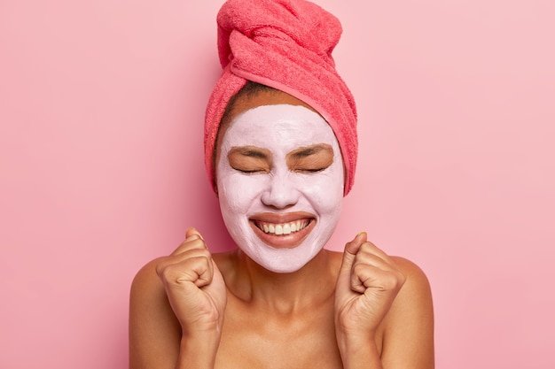 Наполненная энергией счастливая женщина носит глиняную маску на лице, обернутые полотенцем волосы, широко улыбается, сжимает кулаки от удовольствия, изолирована на розовой стене