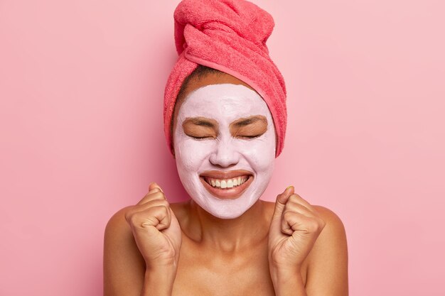 Energized happy woman wears clay mask on face, wrapped towel on hair, smiles broadly, clenches fists from pleasure, isolated over pink wall