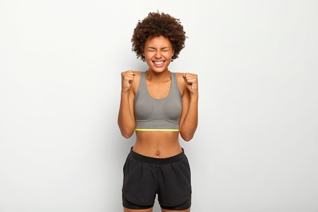 Free photo energized sporty woman rejoices winning, raises clenched fists, smiles broadly, wears sport bra, smiles broadly, isolated over white background,