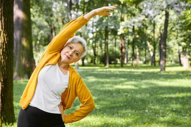 Energy, health, well being and retirement concept. Beautiful sporty senior woman with short hair doing side bend, keeping arm outstretched. Retired female exercising outdoors in park or forest