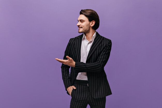 Engaging young brunette-haired boy in light shirt and classic striped suit pointing aside on copy space with one hand isolated over violet wall