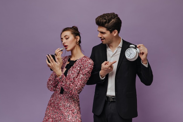 Engaging young man in white shirt and black suit showing time to his girlfriend Lady wearing pink dress making makeup against violet background