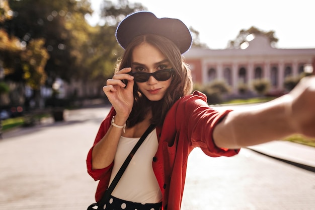 Engaging young Parisienne with dark wavy hair stylish beret and sunglasses in white top and red shirt making selfie against city background Sunny warm weather