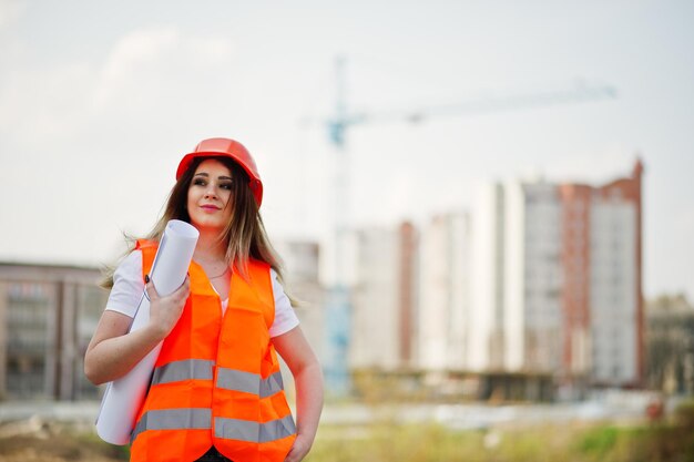 Engineer builder woman in uniform waistcoat and orange protective helmet hold business paper against new buildings with crane