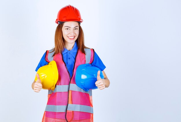 Девушка-инженер в форме держит синие и желтые каски и показывает палец вверх.