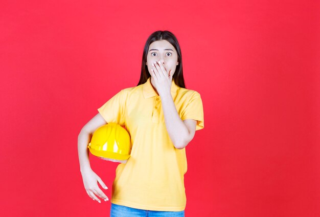 黄色の安全ヘルメットを保持し、恐怖と恐怖に見える黄色のドレスコードのエンジニアの女の子。