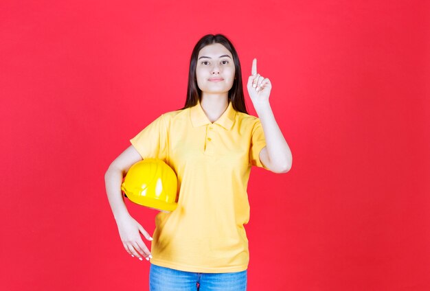Девушка-инженер в желтом дресс-коде держит желтый защитный шлем и где-то показывает