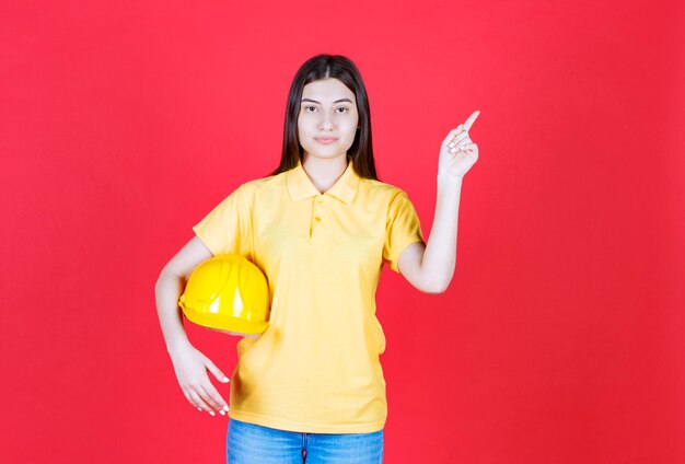 Engineer girl in yellow dresscode holding a yellow safety helmet and showing somewhere