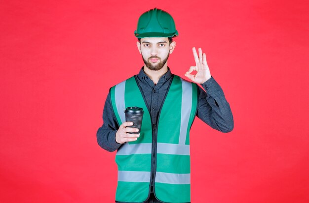 Engineer in green uniform and helmet holding a black disposable cup of coffee and enjoying the taste. 