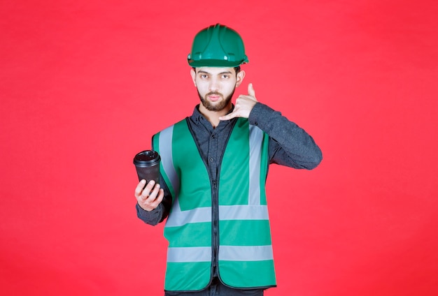 Инженер в зеленой форме и шлеме держит черную одноразовую чашку кофе.