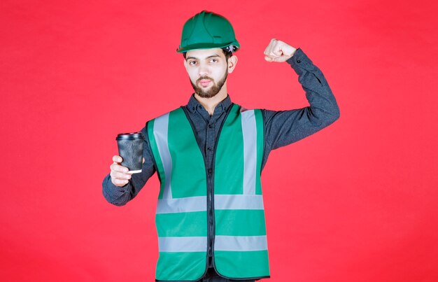 Engineer in green uniform and helmet holding a black disposable cup of coffee. 