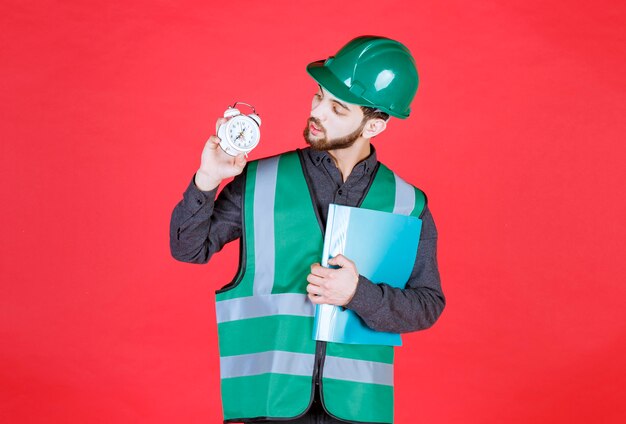 Engineer in green uniform and helmet holding a blue folder and an alarm clock. 